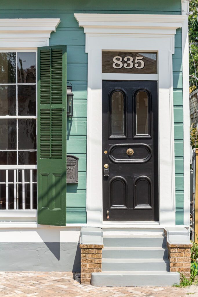Photograph of front of a green shotgun house with a historic wood door with molding painted black, windows and shutters.